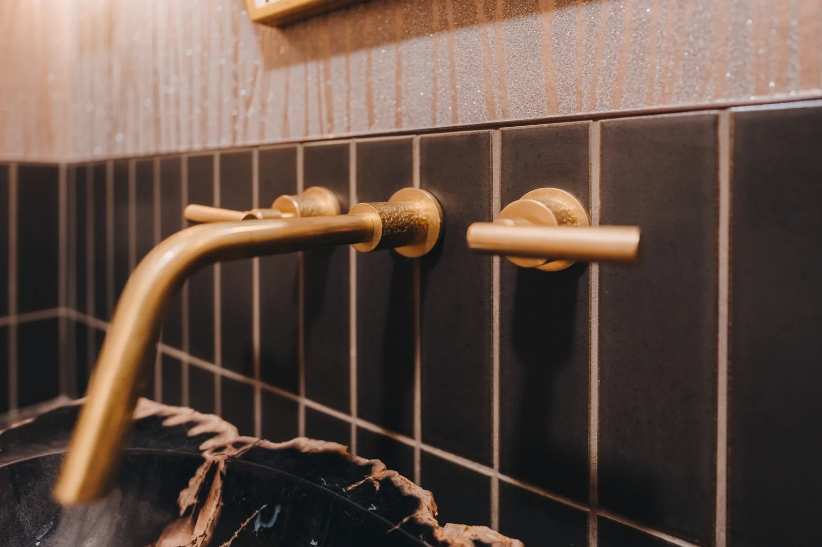 Unlacquered brass wall-mount faucet against dark stacked tile