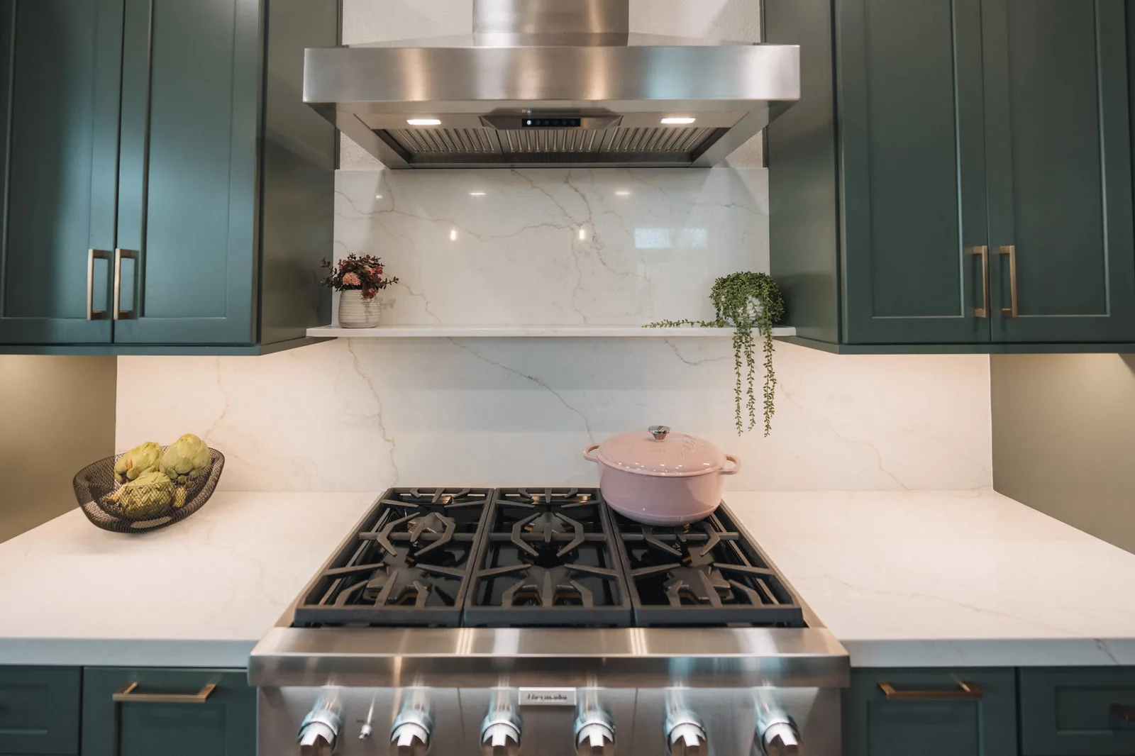 Deep green kitchen cabinets around a stainless range and quartz backsplash